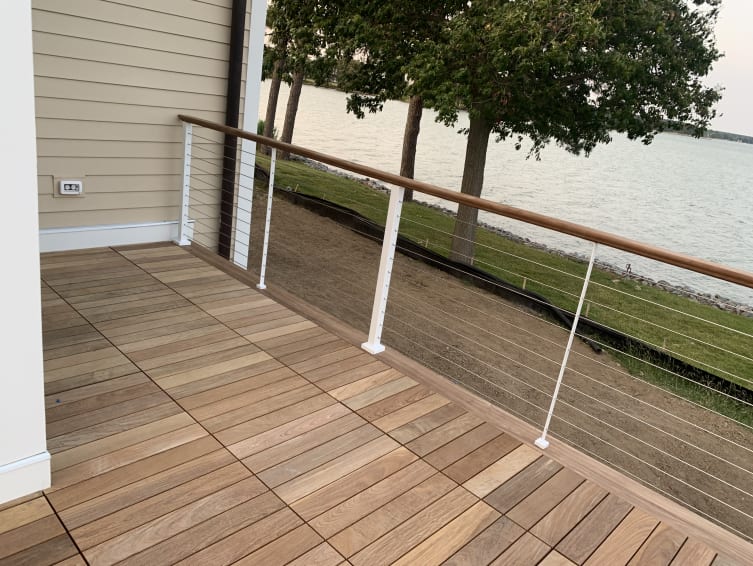A hardwood deck corner with white cable railing posts and wood cap rail overlooking a lake.