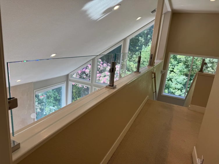 Stainless square posts hold glass panels along a loft knee wall beside angled windows and greenery.