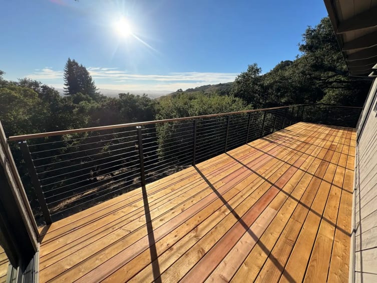 A fresh wood deck with black cable railing and wood cap overlooks wooded hills under a bright sun.