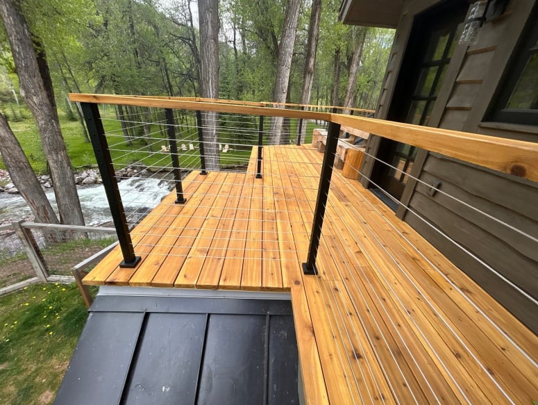Cedar deck on a dark-sided cabin with black cable railing posts and a cedar cap rail overlooking a creek.