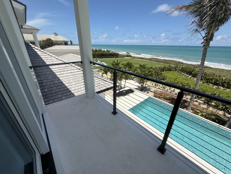 Black cable railing on a covered balcony overlooks a lap pool, palms and the ocean.