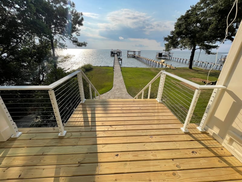 White posts with stainless cable frame a deck stair leading down a lawn to a long dock on the bay.