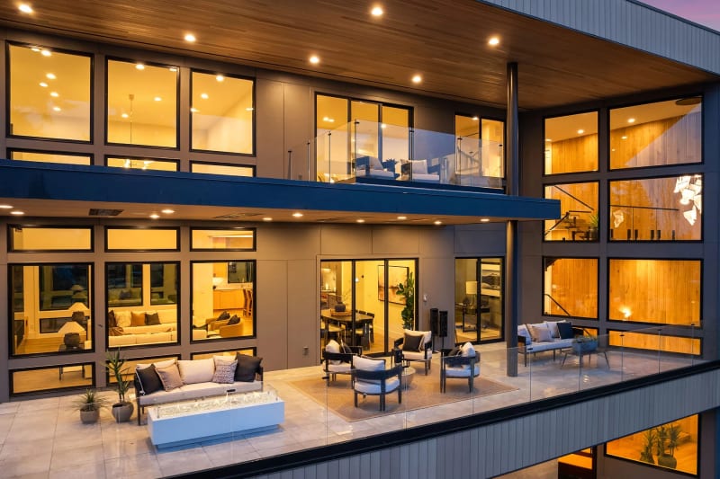 Two-story modern home at dusk with frameless glass railings on the upper balcony and terrace overlooking a lit patio lounge.