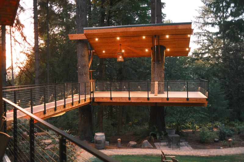 A timber treehouse platform and bridge wrapped in black cable railing glow at dusk among tall firs.