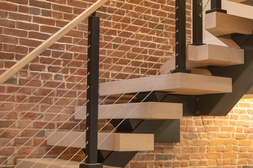 Light maple floating treads on a black mono stringer with cable railing against an exposed brick wall.
