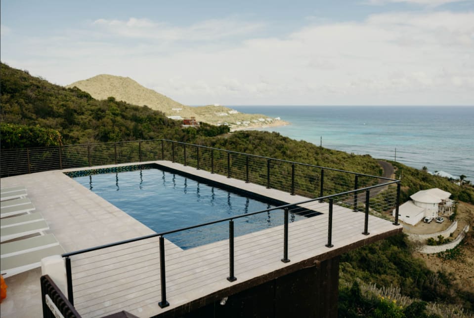 Black surface-mount rod railing with an LED handrail rings a pool deck high above turquoise ocean and green hills.